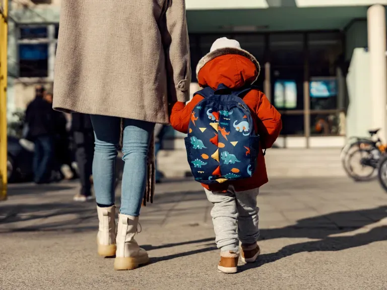 Eine Mutter, die ihr Kleinkind an der Hand hält und in Richtung eines Eingangs eines Gebäudes zugeht.