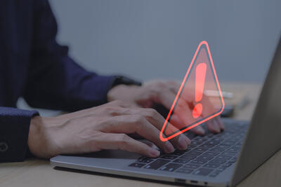 Ein Nutzer tippt an seinem Laptop, darüber schwebt ein rotes Ausrufezeichen.