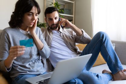 Ein Paar das auf ein Laptop blickt. Die Frau hält eine Kreditkarte in der Hand, der Mann telefoniert